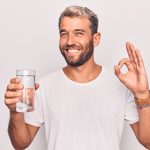 Handsome blond man with beard drinking glass of water to refreshment over white background doing ok sign with fingers, smiling friendly gesturing excellent symbol